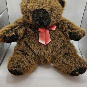 May include: A plush teddy bear with shaggy brown fur, a dark brown snout, and dark brown paws. The bear features a smooth, satin-like pink ribbon tied in a bow around its neck. The toy has rounded ears and a soft, rounded body shape.