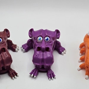 Peut inclure: Trois figurines de jouets colorées : une créature marron semblable à un hippopotame, une créature violette semblable à un hippopotame et une créature orange semblable à un insecte. Chacune a de grands yeux et des accents blancs sur les pieds et les cornes.