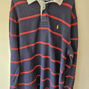 May include: A long-sleeve rugby shirt featuring horizontal red stripes on a navy blue background. The shirt has a light gray collar, a buttoned placket, and a yellow embroidered logo on the left chest. The interior label reads Polo Ralph Lauren.
