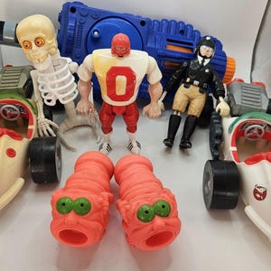 May include: A collection of plastic toys including two white and red vehicles with Ghostbusters logos, a skeleton figure, a red and white wrestler figure, a black and tan police figure, two orange textured creatures, and a large blue plastic blaster.