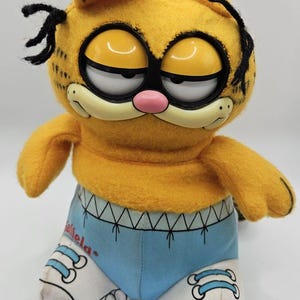 May include: A plush Garfield toy. The orange cat has a sleepy expression with yellow eyes, a pink nose, and black whiskers. He wears blue shorts and white trainers with the word "Garfield" on the side.
