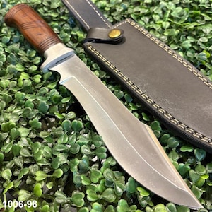 May include: A fixed-blade knife with a polished silver steel blade and a contoured brown wooden handle featuring horizontal grooves. The knife is accompanied by a dark brown leather sheath with visible light-coloured stitching and a brass snap.