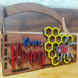 Könnte beinhalten: Ein hölzerner Honigträger mit Griff, der ein Wabenmuster und eine Biene mit gelben, orangefarbenen und roten Akzenten aufweist. Der Träger ist mit "Pure Honey" beschriftet.