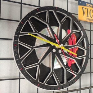 May include: A black and silver clock with a unique design resembling a car wheel. The clock face is made of a CD with a yellow hour hand and a red minute hand. The clock is mounted on a black wire grid.