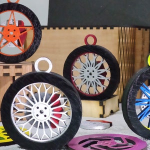 May include: A collection of wooden wheel ornaments with black tires and colorful inner designs. The ornaments feature various spoke patterns and colors, including orange, white, blue, and yellow. Each ornament has a small loop for hanging.