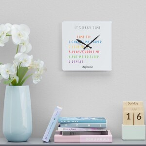 May include: White square clock with black hands and a colourful list of baby care tasks: Change diaper, feed me, play/cuddle me, put me to sleep, repeat. The clock is personalised with the name Stephanie.