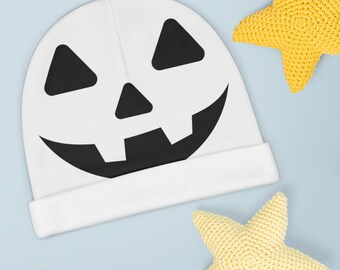 May include: White knitted hat with a black jack-o'-lantern face design. The hat is on a light blue background with two yellow crocheted stars.