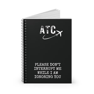 Puede incluir: Cuaderno negro de espiral con el texto "ATC" y un avión blanco. El texto debajo dice "Por favor, no me interrumpas mientras te estoy ignorando."