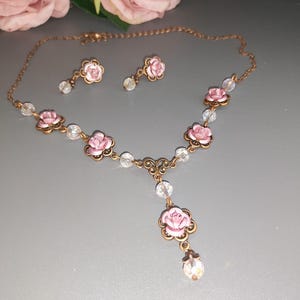 May include: A vintage-style jewelry set featuring a necklace and matching earrings. The set includes a delicate chain, pink rose-shaped pendants, and clear crystal accents. The necklace has a drop pendant with a single crystal. The earrings have a single crystal drop.