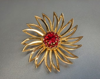 Op de afbeelding: Goudkleurige bloembroche met een levendig rood centrum. De broche heeft een gedetailleerd ontwerp met opengewerkte bloemblaadjes die uitstralen vanuit een centrale cluster van rode stenen. Een klassiek sieraad.