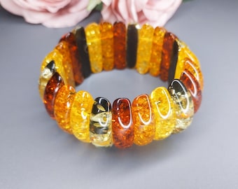 Könnte beinhalten: Ein elastisches Armband aus ovalen, bernsteinfarbenen Perlen. Die Perlen variieren in den Farbtönen Gelb, Orange und Braun und erzeugen eine warme, natürliche Farbpalette. Das Armband liegt auf einer grauen Oberfläche, im Hintergrund ist eine verschwommene rosa Blume zu sehen.