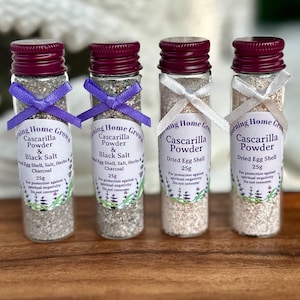 May include: Four small glass bottles with white lids and purple ribbons. Each bottle contains a white powder and has a label that reads "Cascarilla Powder" and includes a description of the contents. The bottles are arranged in a row on a wooden surface.