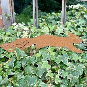 May include: A rusty brown metal squirrel silhouette, running across a bed of green and white ivy. The squirrel has cut-out details for its eye and tail. The background includes a brick wall and a metal fence.