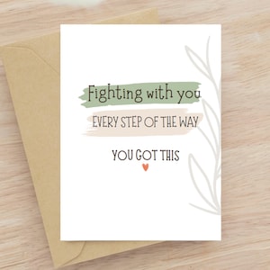 Op de afbeelding: Een witte wenskaart met de tekst "Fighting with you every step of the way, you got this" in zwarte letters. De kaart heeft een groene en bruine achtergrond met een klein rood hart. De kaart zit in een bruine envelop.