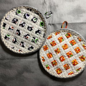 May include: Two round quilted pot holders. One features a pattern of various eye designs in black, white, and green. The other has a pattern of orange, yellow, and white candy corn. Both have gray borders and hanging loops.