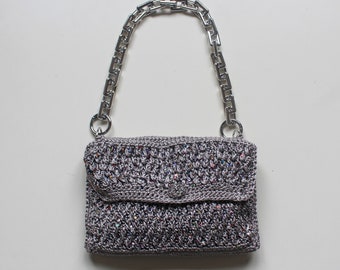 May include: A silver chain strap handbag with a gray crocheted body. The bag has a flap closure and a small silver button.