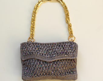 May include: A small gray crocheted handbag with a gold chain strap. The bag has a sparkly texture and a flap closure.