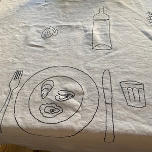 May include: A white tablecloth with black embroidery of a dinner setting, including a plate of oysters, a glass, a bottle, a fork, and a knife.
