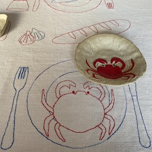 May include: White tablecloth with embroidered red and blue food illustrations, including a crab, fish, bread, and shells. A small, round ceramic bowl with a red crab design sits on the cloth.
