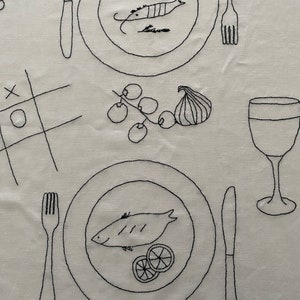 May include: A white tablecloth with black embroidery depicting a simple table setting with two plates, forks, knives, a glass of wine, and a branch of grapes with a fig. The plates have fish and lemon slices embroidered on them.