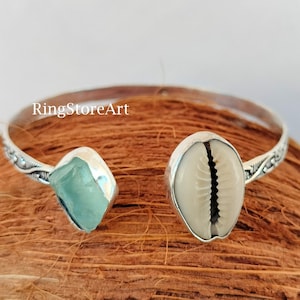 May include: Silver bangle bracelet with a light blue stone and a cowrie shell. The bracelet has a decorative design on the band.