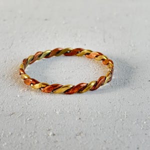Może przedstawiać: Okrągła bransoletka typu bangle wykonana ze skręconych pasm metalicznej miedzi i mosiądzu. Wzór charakteryzuje się powtarzalnym układem naprzemiennych segmentów z ciepłej czerwonobrązowej miedzi oraz jasnego żółtego mosiądzu, tworząc ciągłą plecioną fakturę.