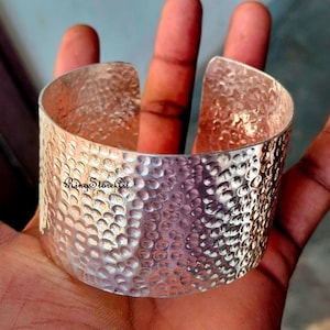 Puede incluir: Un brazalete ancho de plata con una textura martillada. El brazalete tiene forma curva y está abierto en la parte superior. El texto "Ring Store Art" es visible en el brazalete.