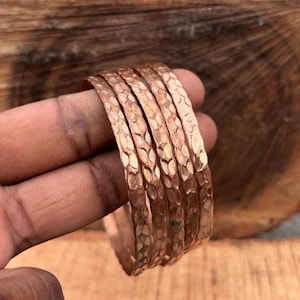 May include: A set of five copper bangles featuring a hammered texture across the outer surface. Each bangle has a consistent metallic reddish-brown finish and a rounded profile, creating a stackable design that reflects light across the indentations.