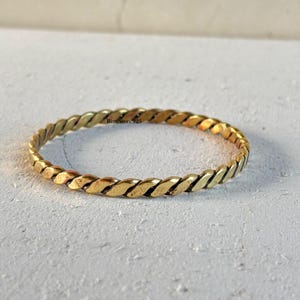 Może przedstawiać: Złota metalowa bransoletka typu bangle z ciągłym wzorem skręconej liny. Powierzchnia metalu ma polerowane wykończenie z subtelnymi ciemnymi akcentami w rowkach splotu. Na obręczy widoczny jest napis RingStore925.