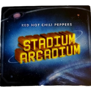 May include: CD case for the Red Hot Chili Peppers album "Stadium Arcadium." The cover features the album title in 3D yellow letters against a blue and black space-themed background with a planet and stars. The band's name is at the top.