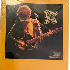 May include: A sealed compact disc featuring a cover photograph of a musician playing an acoustic guitar and a harmonica on a stand. The text Real Live is printed in a stylized yellow font in the upper right corner, with the compact disc logo below.