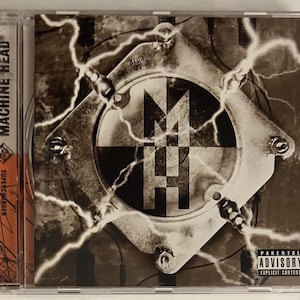 May include: A CD case with a black and white image of a machine head with lightning bolts. The text "Supercharger" and "Machine Head" are visible on the case. The back of the case has a Parental Advisory sticker.