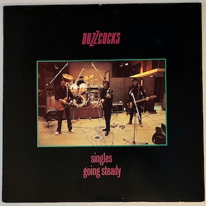 May include: A black and white photo of a band, the Buzzcocks, in a recording studio. The band members are standing in front of a microphone and a drum set. The text "singles going steady" is printed below the image.