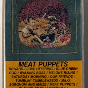 May include: A cassette tape case with a yellow label featuring the band Meat Puppets. The case has a green background with a photo of a baby sitting on a wooden platform surrounded by flowers. The text on the label reads "Meat Puppets" and lists the songs on the cassette tape: "Reward", "Love Offering", "Blue-Green", "God", "Walking Boss", "Melons Rising", "Saturday Morning", "Our Friends", "Tumblin' Tumbleweeds", "Milo, Sorghum and Maize", "Meat Puppets", "Playing Dead", "Litterbox", "Electromud", "The Gold Mine". The label also includes the cassette tape's catalog number, "SSTC 009", the type of tape, "CrO2", the running time, "TT-20:52", and the price, "$6.98".