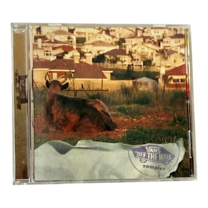 May include: A CD case featuring a photograph of a deer lying in a field with a suburban backdrop. The bottom right corner displays a white and blue Vans Off The Wall sampler logo.