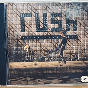 May include: A CD case with a photo of a young person kicking a skull in front of a wall with the word "CUE" written in large letters. The text "ROLL THE BONES" is written below the word "CUE". The CD case has a "FINYL" sticker on the bottom right corner.