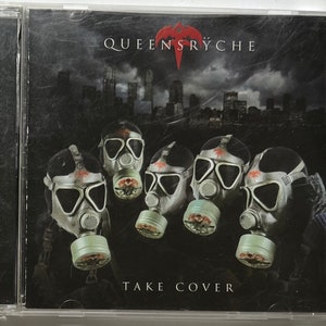 May include: A CD case featuring the band Queensrÿche. The album cover shows five figures wearing gas masks against a dark, apocalyptic cityscape. The band's name and the album title, "Take Cover," are displayed.