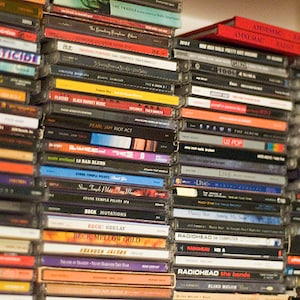 May include: A close-up view of a shelf filled with a variety of compact discs (CDs). The CDs are stacked vertically and horizontally, with the spines facing outward. The CDs are a mix of different colors, including black, silver, gold, and red. Some of the CDs have text on the spines, including the names of artists and albums. The CDs are arranged in a random order, creating a visually interesting pattern.