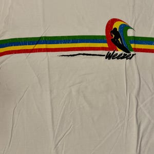 May include: White t-shirt featuring a colorful graphic of a surfer riding a wave, with a rainbow arch and the word "Weezer" in black. The design includes horizontal stripes in green, yellow, red, and blue.