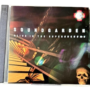 May include: A Soundgarden CD case with the album title "Alive in the Superunknown" in white letters. The cover art features a surreal image with a spider, skull, and geometric shapes in shades of brown, green, and purple.