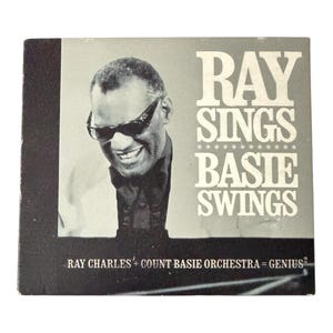 May include: A vintage album cover for "Ray Sings, Basie Swings" featuring a black and white image of Ray Charles wearing sunglasses. The album title is in large white letters, with the text "Ray Charles + Count Basie Orchestra = Genius²" below.