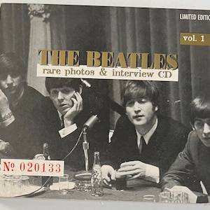 May include: A black and white photo of The Beatles on a CD cover. The cover is gold with the text "THE BEATLES rare photos & interview CD" and "LIMITED EDITION vol. 1". The photo shows the four band members sitting at a table with microphones and drinks in front of them. The number "N° 020133" is printed in red on the bottom left corner of the cover.