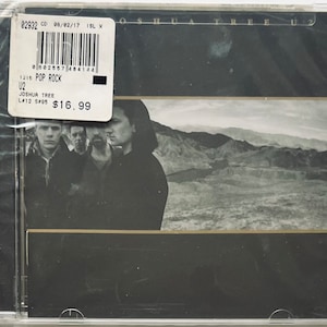 May include: A CD case with a black and white photo of a band on the front. The band is standing in front of a desert landscape. The title of the album is "Joshua Tree" and the band is U2. The CD is priced at $16.99.