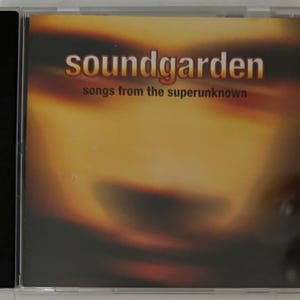 May include: A Soundgarden CD case with the album title "songs from the superunknown" in red text. The band name "soundgarden" is in white and orange text. The background is a blurred orange and brown gradient.