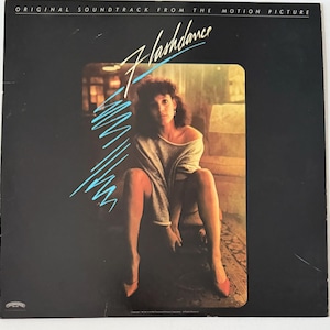 May include: A vintage vinyl record album cover featuring a woman in a grey shirt sitting on a chair. The album cover is for the soundtrack to the movie "Flashdance".