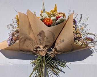May include: A bouquet of dried flowers wrapped in brown paper with the text The Flower Patch printed on the front. The arrangement features a mix of orange, yellow, white, and purple blossoms with green stems tied together with twine.
