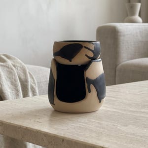 Puede incluir: Un calentador de cera de cerámica de dos piezas con una base de color beige mate y patrones de goteo pintados en negro irregular. La sección superior es un cuenco extraíble con un diseño a juego en negro y beige, con una gran abertura.