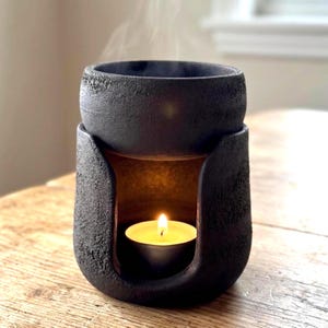 May include: A matte black ceramic oil burner with a rough, textured surface. The cylindrical base features a wide arched opening containing a small lit tea light candle. A bowl sits on top, emitting a thin wisp of smoke from the heated contents.