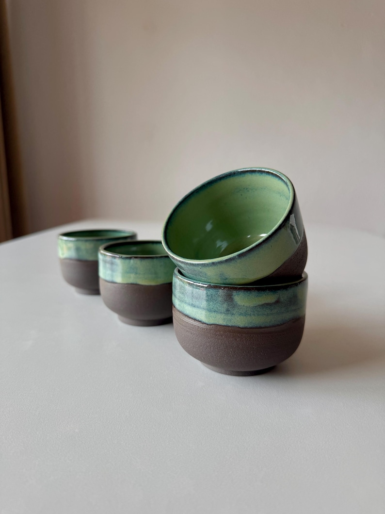 May include: Four small ceramic cups with a green and brown glaze. The cups are stacked on top of each other.
