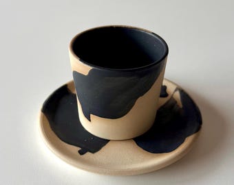 Może przedstawiać: Zestaw filiżanki i spodeczka ceramicznych w kolorze czarnym i beżowym z abstrakcyjnym wzorem. Filiżanka i spodeczek są pokryte czarną i beżową glazurą, a czarna glazura tworzy abstrakcyjny wzór na powierzchni.